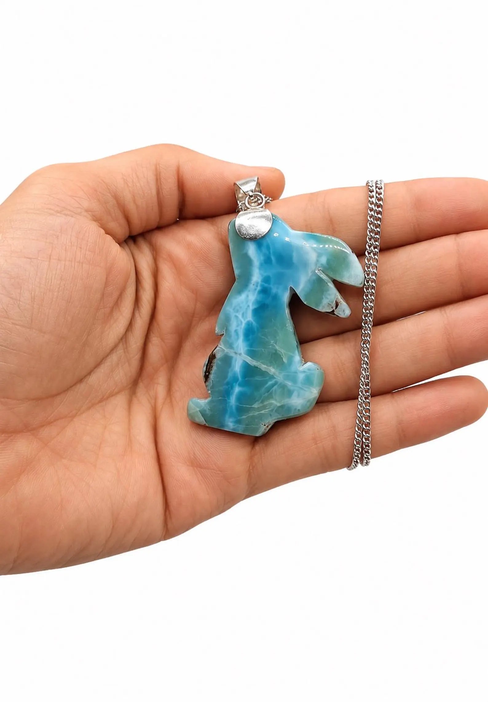 A hand holding a carved blue  Larimar and white marbled pendant on a silver chain, shown against a plain white background.