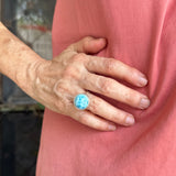 A close-up of a person wearing a textured turquoise ring with a silver-toned band, shown on their ring finger while wearing a pink sleeveless top.