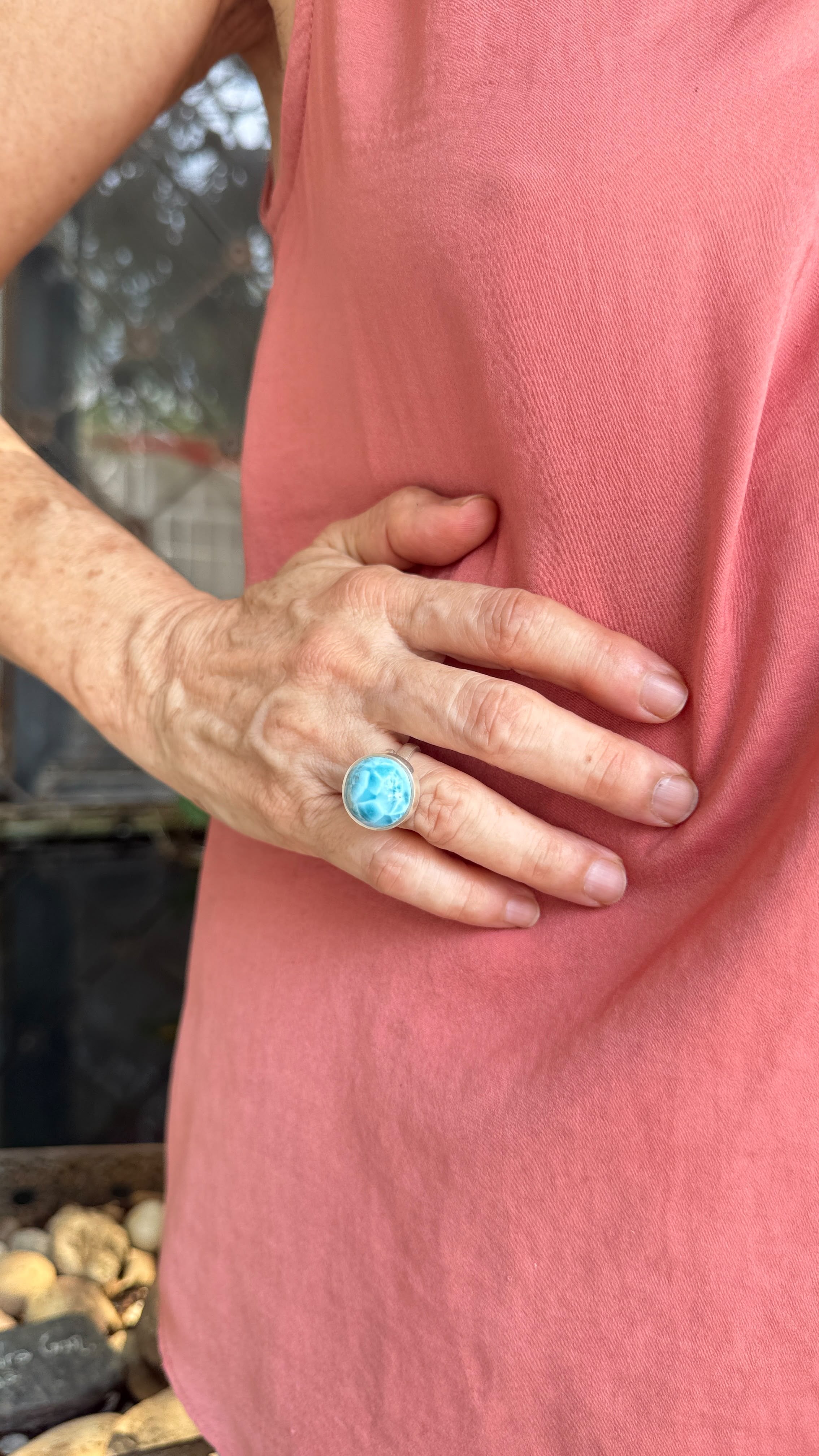 A close-up of a person wearing a textured turquoise ring with a silver-toned band, shown on their ring finger while wearing a pink sleeveless top.