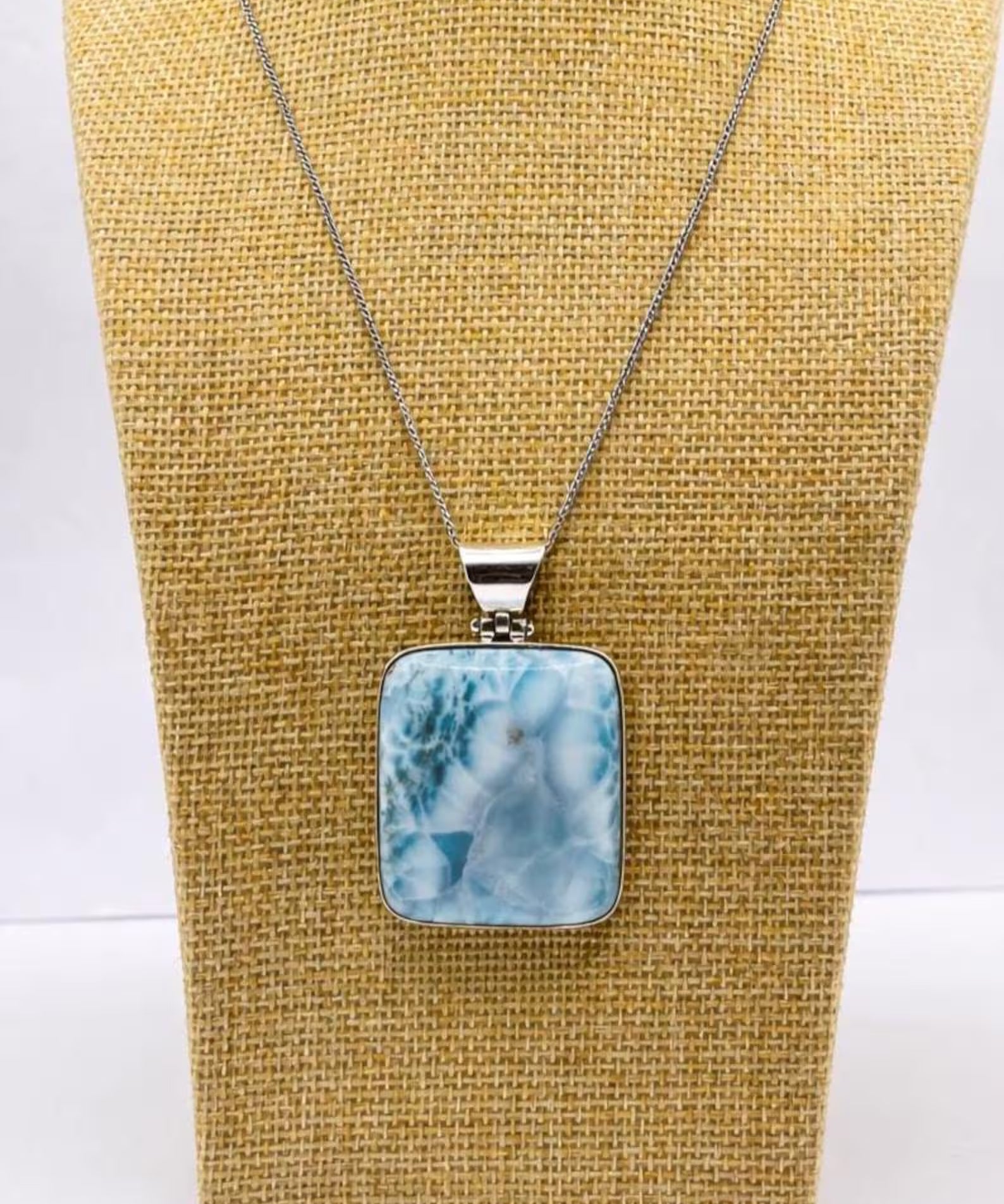 A silver necklace with a square larimar pendant featuring a light blue and white marbled pattern, displayed on a tan woven jewelry bust against a white background.