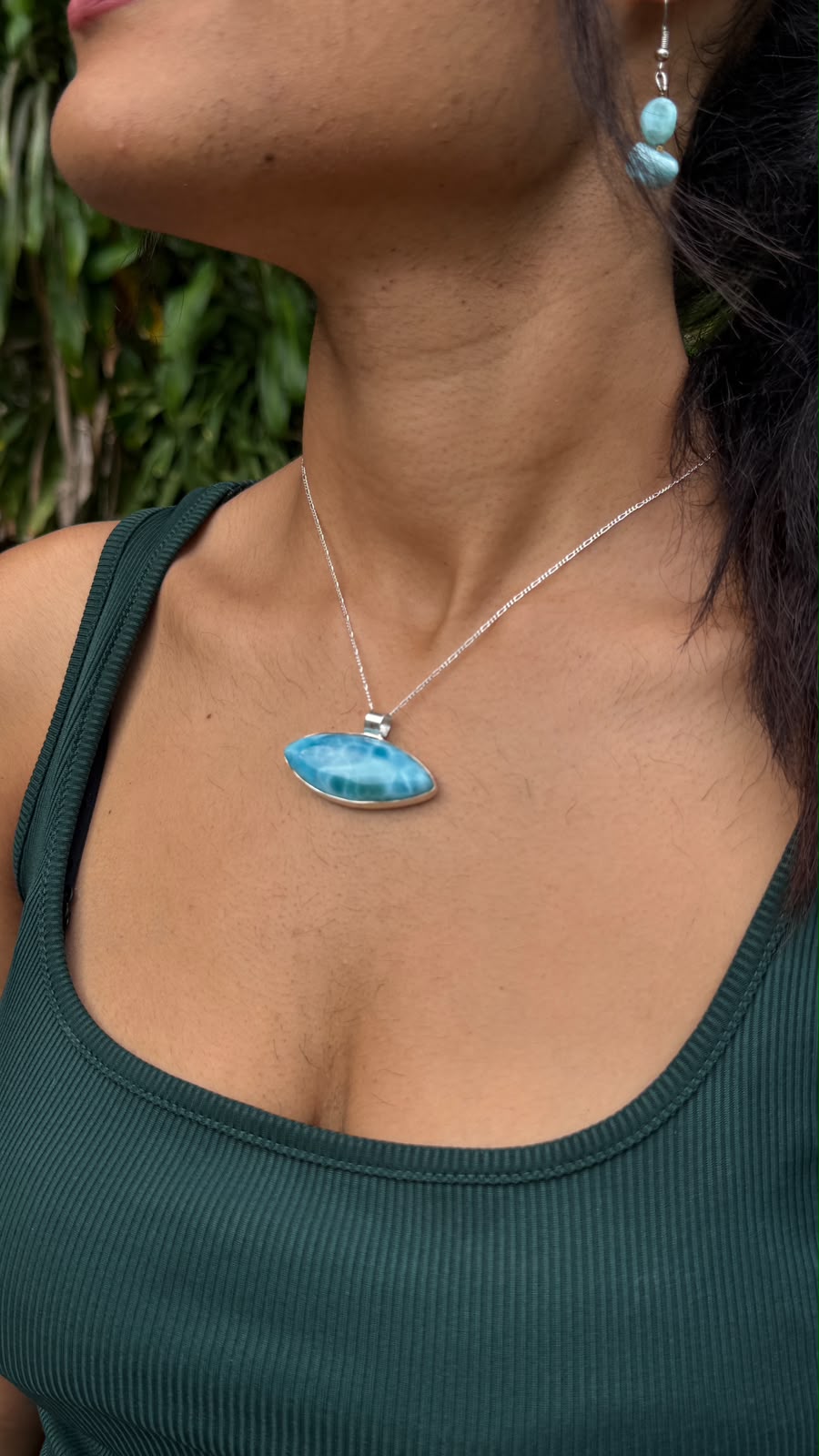 A close-up lifestyle shot of a woman wearing a silver necklace with a vibrant blue marbled labradorite pendant, paired with a dark green ribbed tank top.