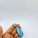 A hand holding a teardrop-shaped blue marbled stone pendant with a small matching blue bead and silver-toned metal settings, against a plain white background.
