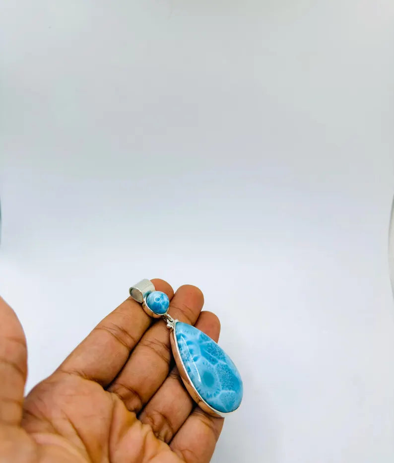 A hand holding a teardrop-shaped blue marbled stone pendant with a small matching blue bead and silver-toned metal settings, against a plain white background.