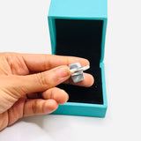 A hand holding a silver-toned ring in front of an open turquoise jewelry box on a white background.