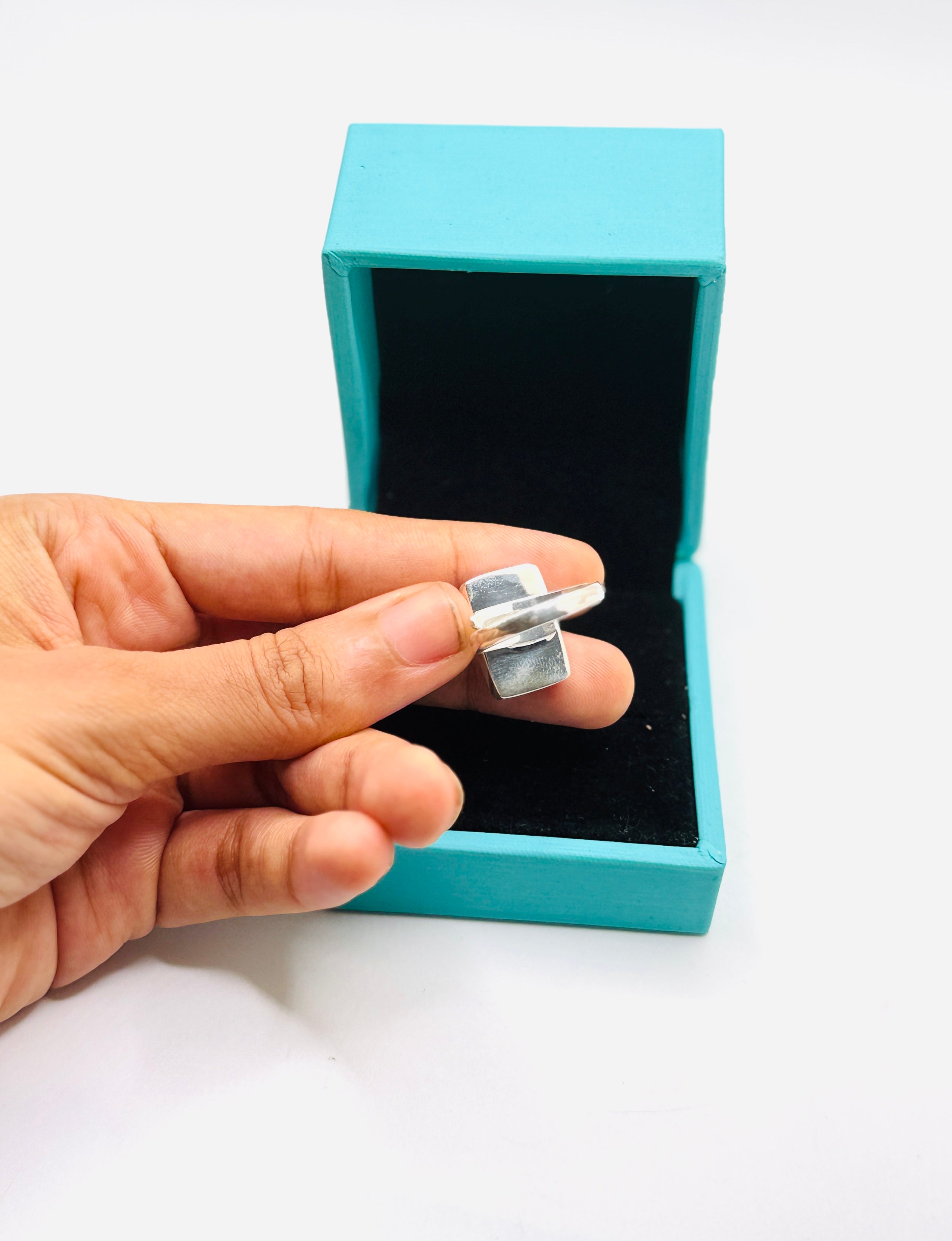 A hand holding a silver-toned ring in front of an open turquoise jewelry box on a white background.