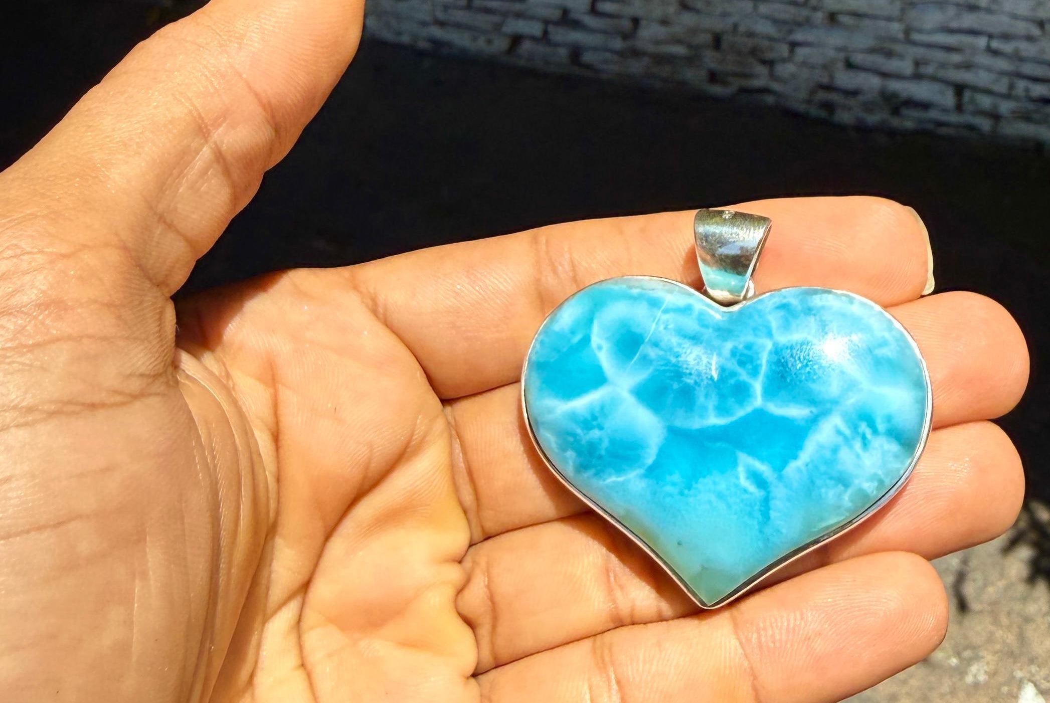A heart-shaped Larimar pendant with a silver bezel, held in a person's hand against an outdoor stone wall background.