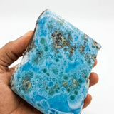 A hand holding a large, Larimar blue stone  specimen with white veining and dark metallic inclusions, set against a plain white background.
