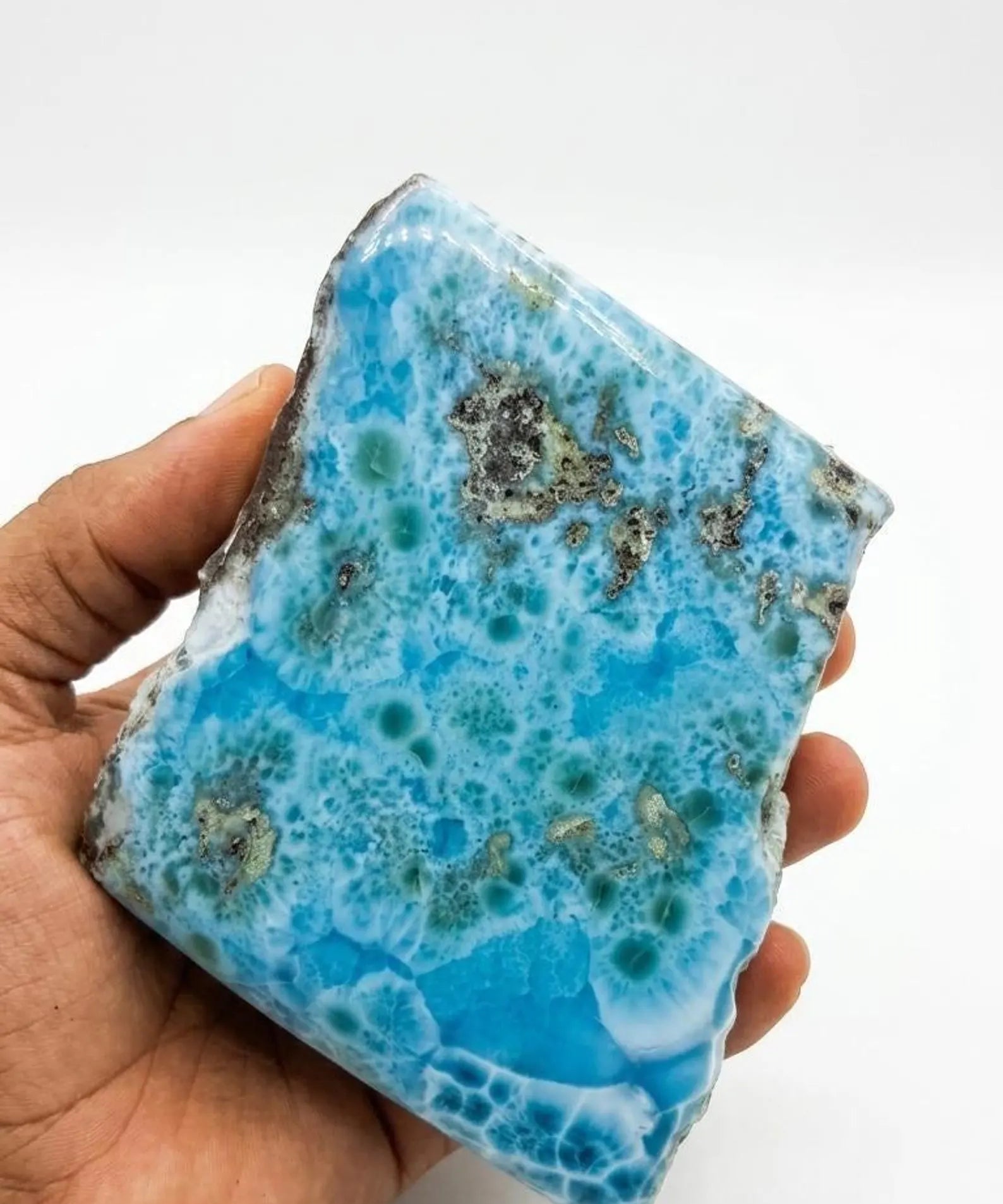 A hand holding a large, Larimar blue stone  specimen with white veining and dark metallic inclusions, set against a plain white background.