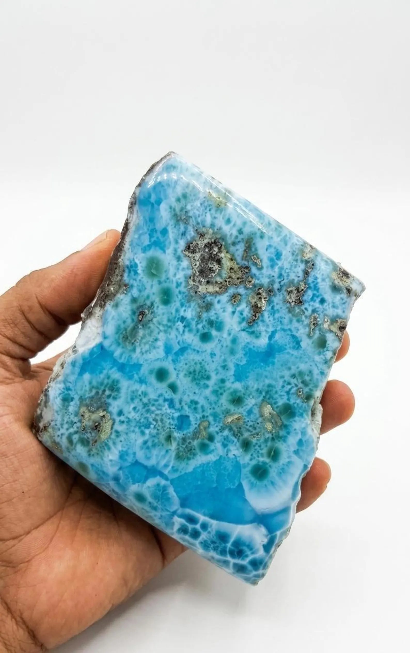 A hand holding a large, Larimar blue stone  specimen with white veining and dark metallic inclusions, set against a plain white background.