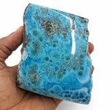 A hand holding a polished blue Larimar stone slab with natural mottled patterns and rough edges against a plain white background.