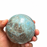A hand holding a polished turquoise-blue and brown stone sphere against a plain white background.