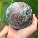 A person's hands holding a round, porous stone sphere with a mottled brown and turquoise patina, set against a background of a large green banana leaf.
