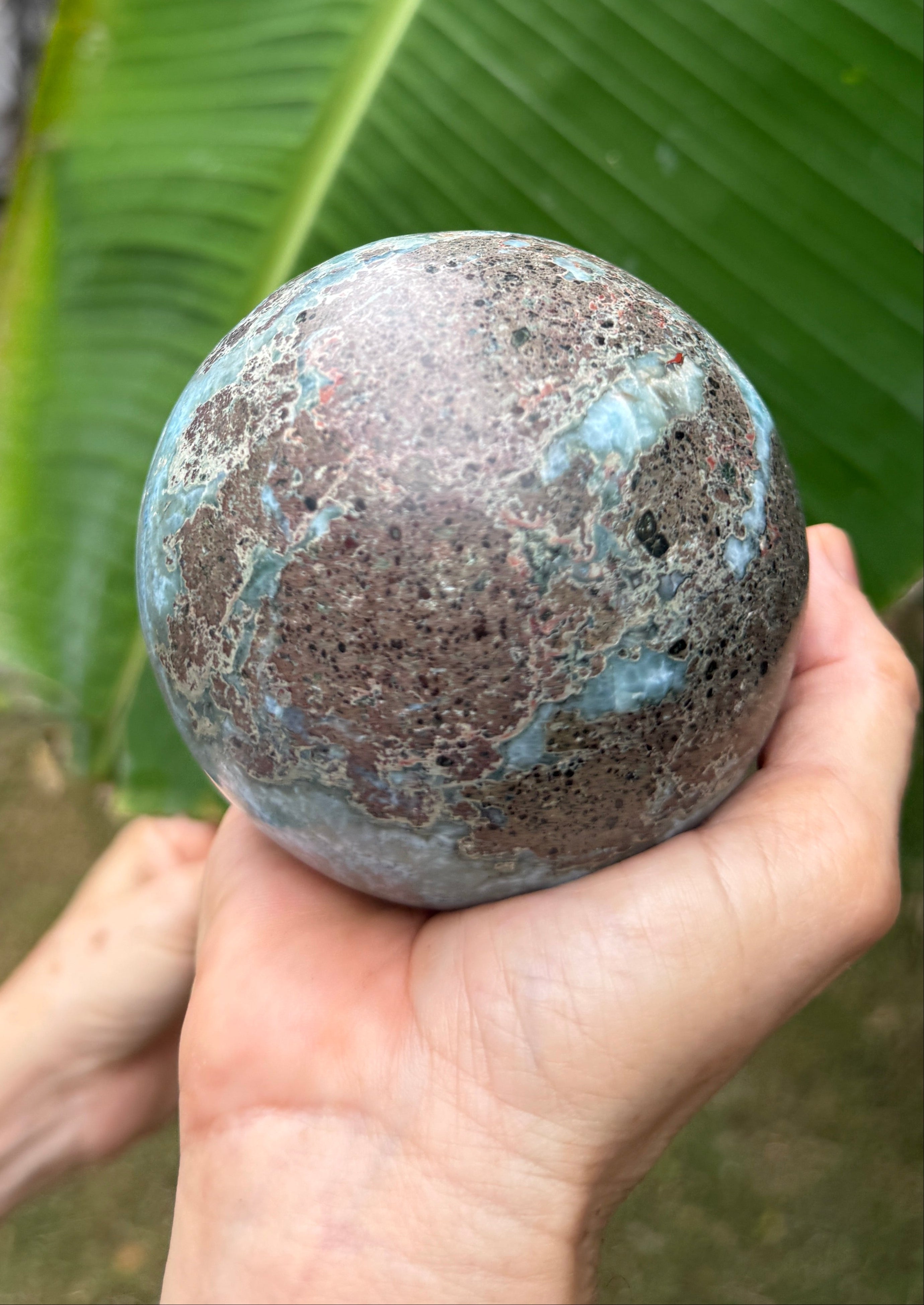 A person's hands holding a round, porous stone sphere with a mottled brown and turquoise patina, set against a background of a large green banana leaf.