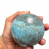 A hand holding a polished blue-green stone sphere against a plain white background.