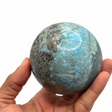 A hand holding a polished larimar stone sphere with natural marbled patterns, shown against a plain white background.