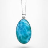 An oval pendant necklace featuring a vibrant blue and turquoise marbled pattern, set in a silver-toned bezel on a delicate silver chain, presented against a clean white background.