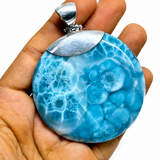 A hand holding a round Larimar pendant with a vibrant blue and white marbled pattern, featuring a polished silver-toned metal cap and bail, set against a plain white background.
