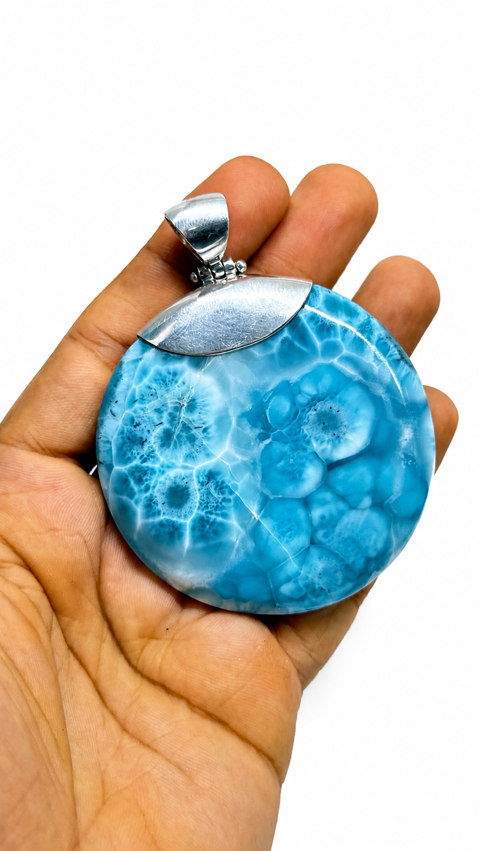 A hand holding a round Larimar pendant with a vibrant blue and white marbled pattern, featuring a polished silver-toned metal cap and bail, set against a plain white background.
