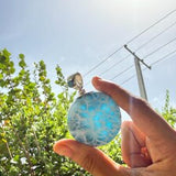 A hand holding a round, light blue marbled stone or glass pendant against a bright outdoor background of green trees, a blue sky, and the sun.