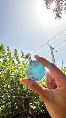 A hand holding a round, light blue marbled stone or glass pendant against a bright outdoor background of green trees, a blue sky, and the sun.